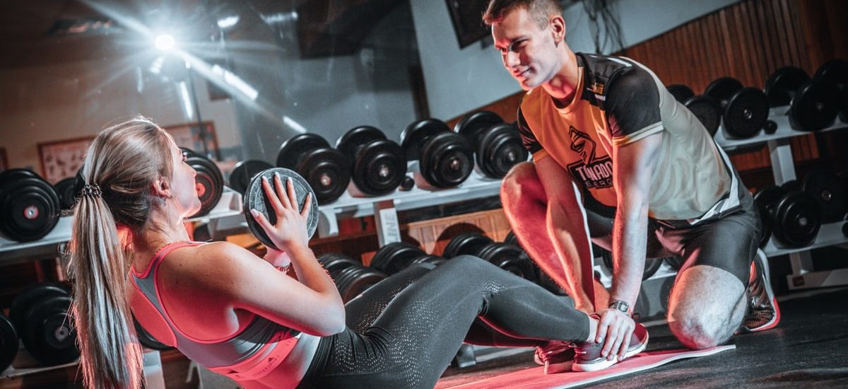 A personal trainer assists a woman doing sit-ups with a medicine ball in a gym setting.