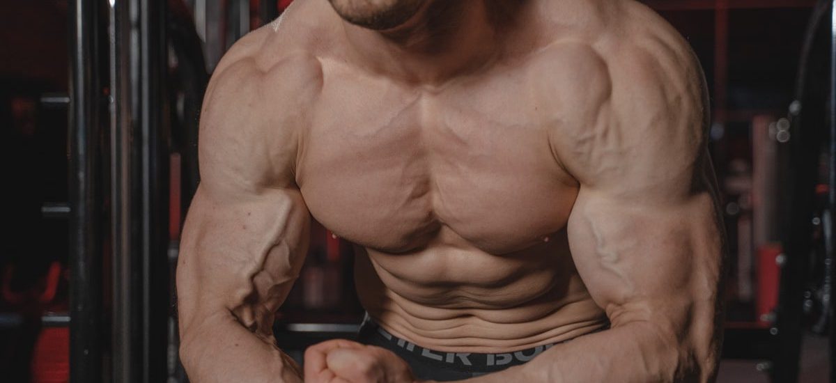 Shirtless man displaying muscular physique with focus on biceps and abs in a gym.