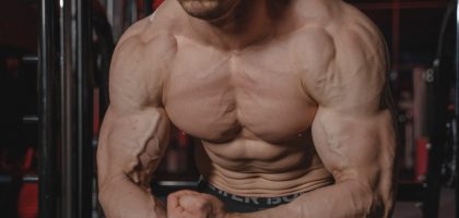 Shirtless man displaying muscular physique with focus on biceps and abs in a gym.