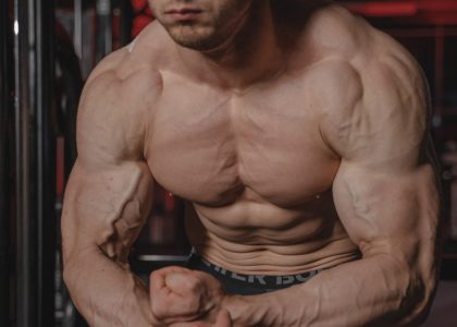 Shirtless man displaying muscular physique with focus on biceps and abs in a gym.